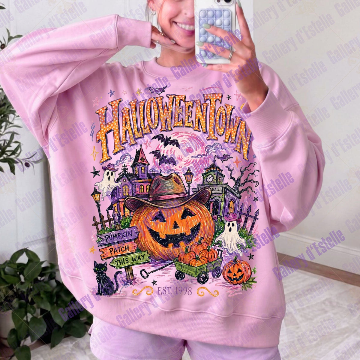 A person taking a mirror selfie wearing a light pink sweatshirt with a large colorful 'Halloween Town' graphic on the front, featuring a jack-o'-lantern, haunted house, pumpkins, ghosts, and bats.