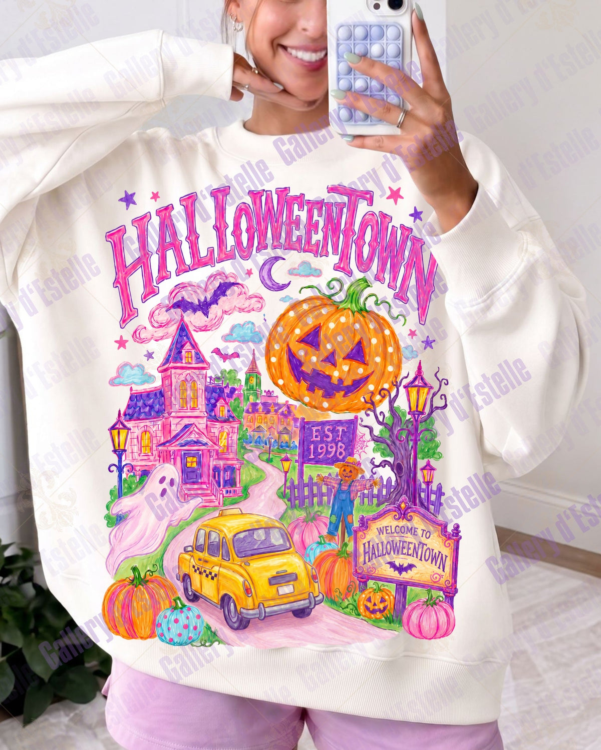 A woman takes a mirror selfie wearing a cream-colored sweatshirt with a colorful 'Halloween Town' graphic print featuring a jack-o'-lantern, haunted house, yellow car, and pumpkins.