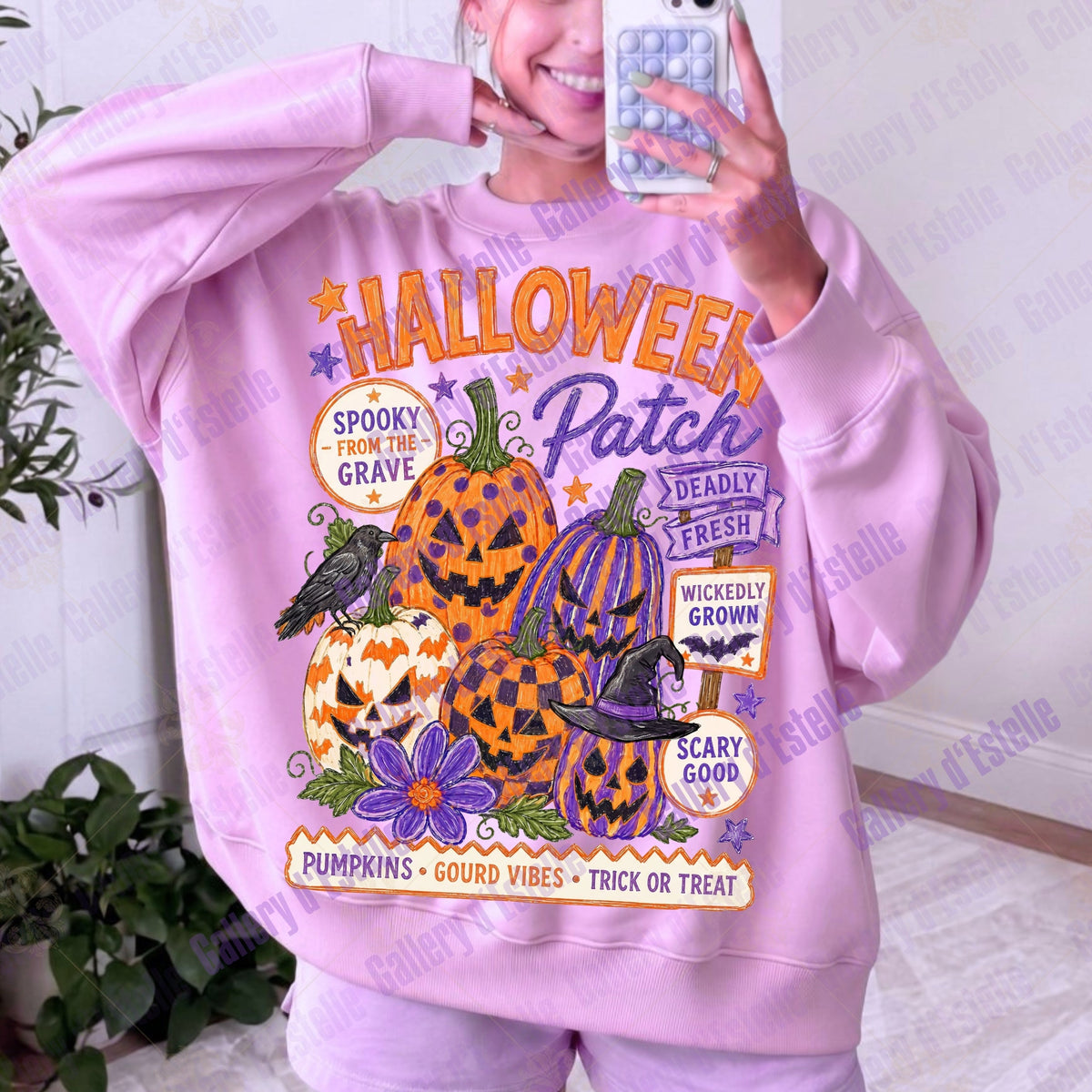 A person taking a mirror selfie wearing a light pink sweatshirt with a large Halloween graphic print featuring pumpkins, a witch hat, bats, and text like 'Halloween Patch' and 'Spooky from the Grave'.