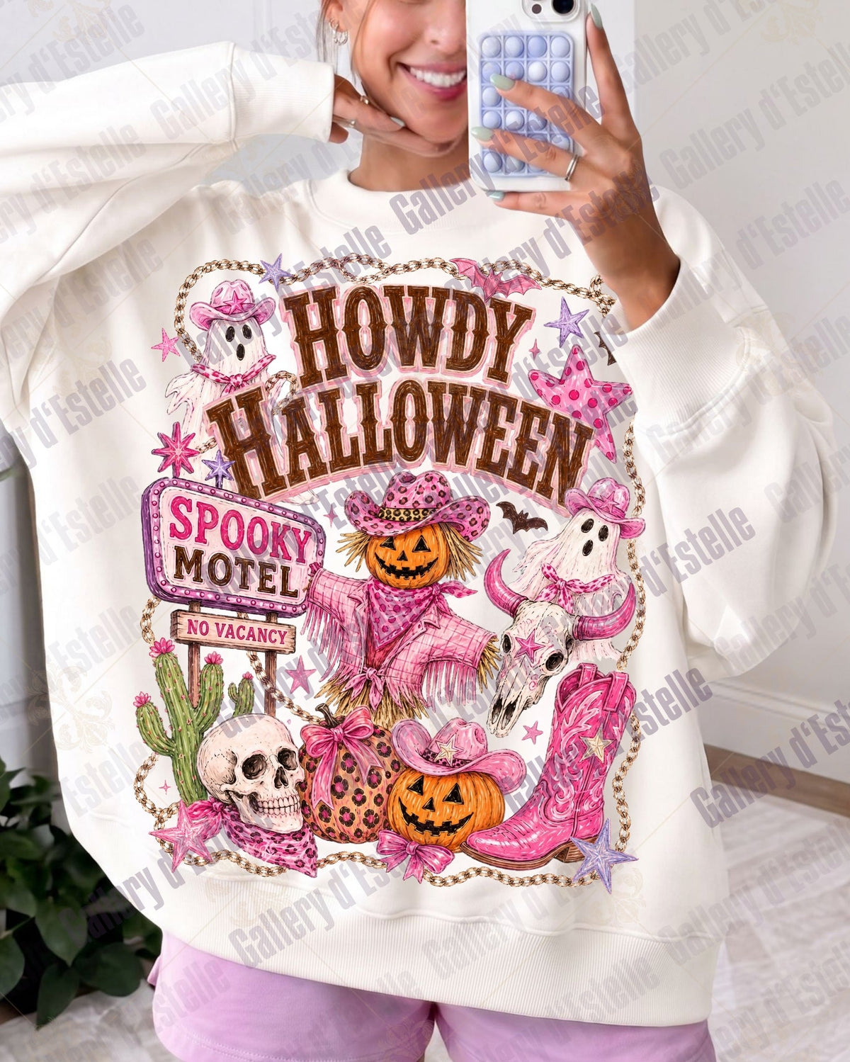 A woman takes a mirror selfie wearing a cream-colored sweatshirt with a large, colorful 'Howdy Halloween' graphic on the front. The graphic features pink ghosts, jack-o'-lanterns, skulls, and a 'Spooky Motel' sign.