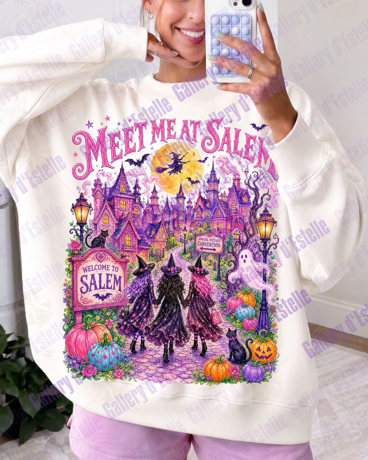 A woman takes a mirror selfie wearing a cream-colored sweatshirt with a large, colorful 'Meet Me at Salem' Halloween graphic on the front, paired with light purple pants.