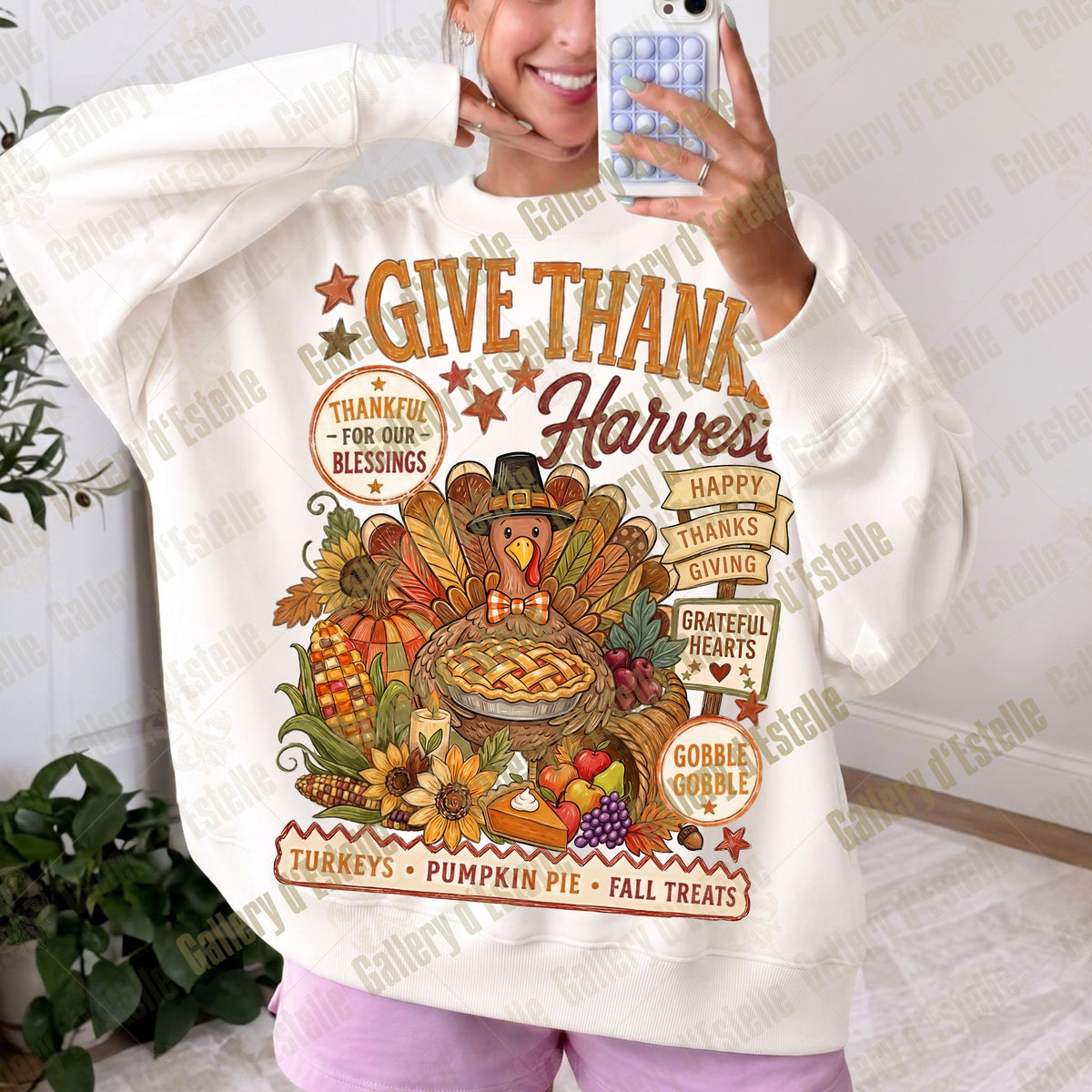 A woman takes a mirror selfie wearing a cream-colored hoodie with a large, colorful Thanksgiving graphic on the front. The graphic features a turkey, a pumpkin pie, autumn leaves, and text reading 'GIVE THANK Harvest' and 'THANKFUL FOR OUR BLESSINGS'.