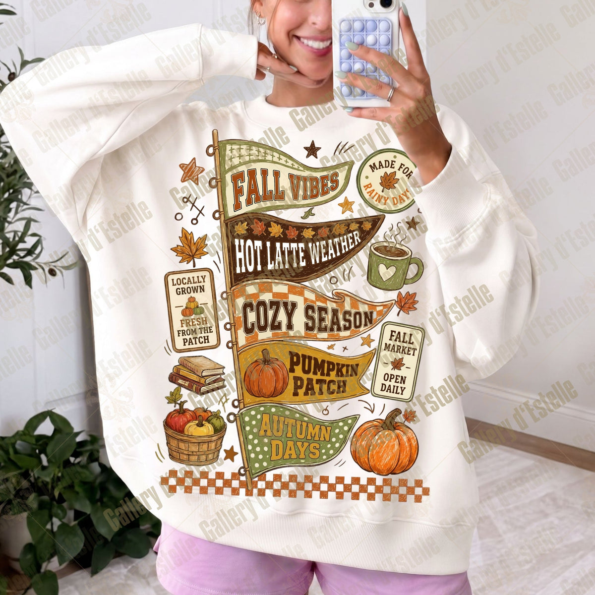 A woman takes a mirror selfie wearing an oversized cream sweatshirt with a detailed autumn-themed graphic print featuring pumpkins, fall leaves, and cozy season text.