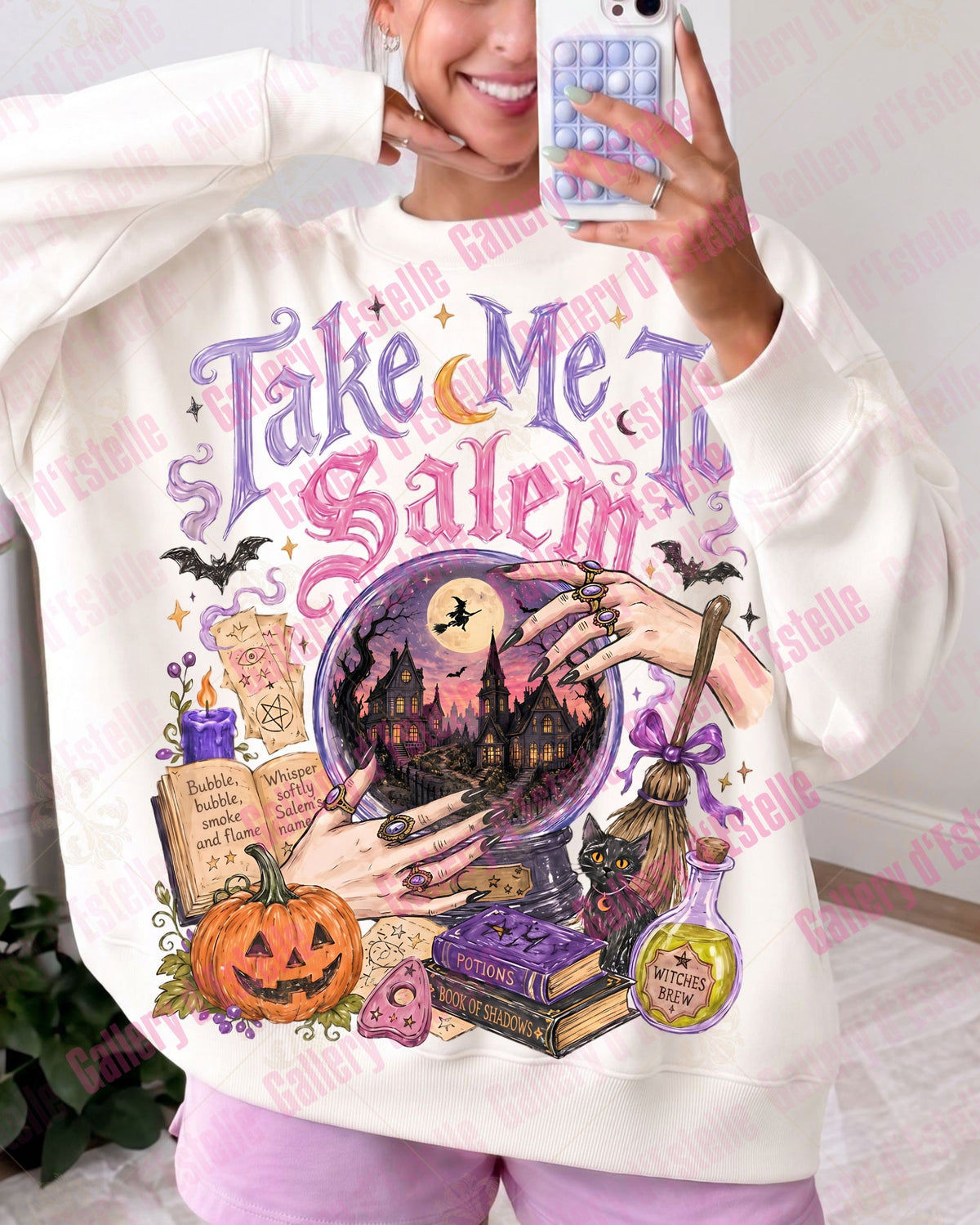A woman takes a mirror selfie wearing a cream-colored sweatshirt with a large, colorful Halloween-themed graphic that reads 'Take Me To Salem' above a spooky illustration of a pumpkin, broom, potion, and haunted house. She is wearing pink pants in a home setting.