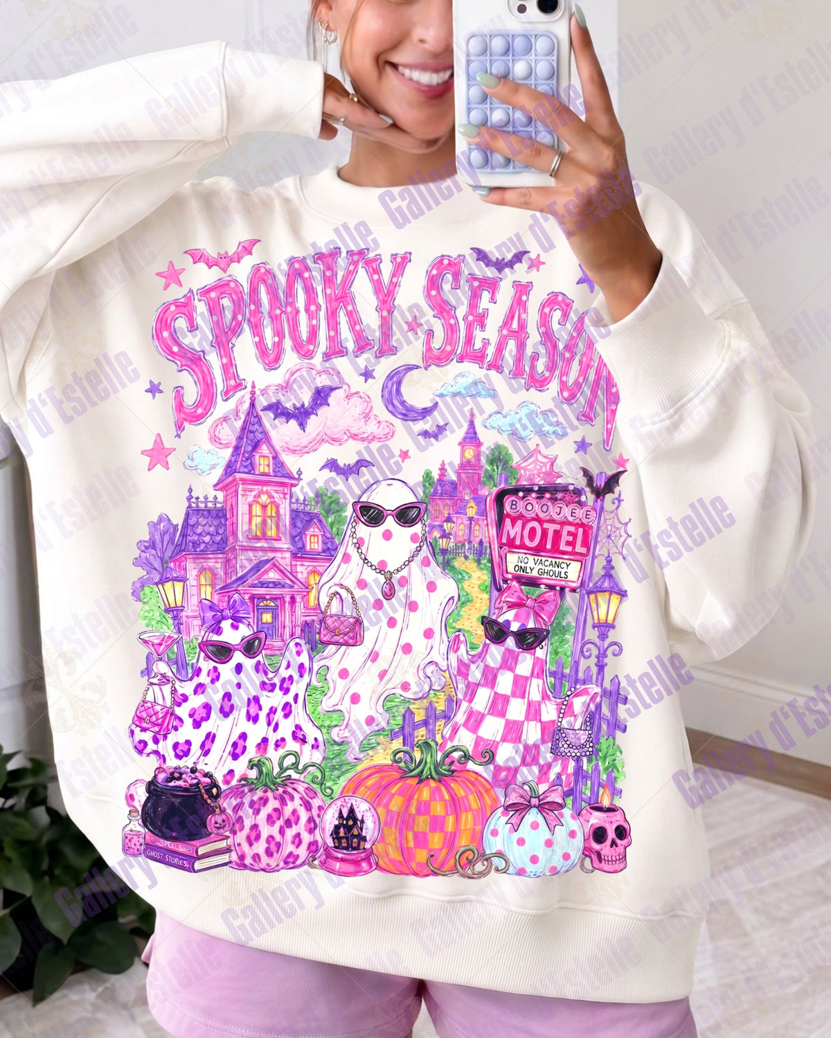 A woman takes a mirror selfie wearing an oversized cream-colored sweatshirt with a colorful 'Spooky Season' graphic print featuring ghosts, pumpkins, a motel sign, and purple leopard print details.
