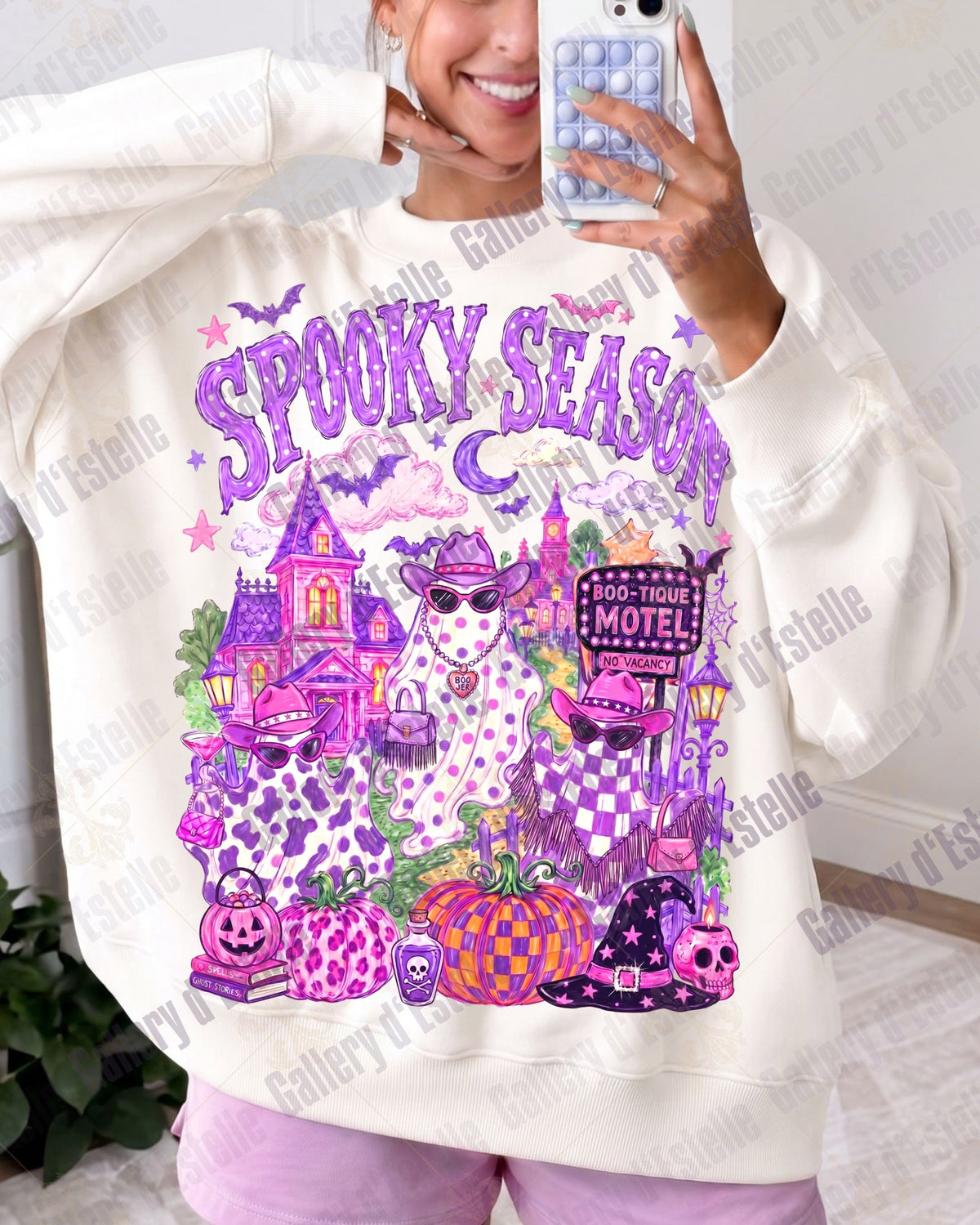 A woman takes a mirror selfie wearing a cream-colored sweatshirt with a large, colorful 'Spooky Season' graphic on the front. The graphic features purple and pink Halloween-themed illustrations including a haunted house, ghosts, pumpkins, and a motel sign.