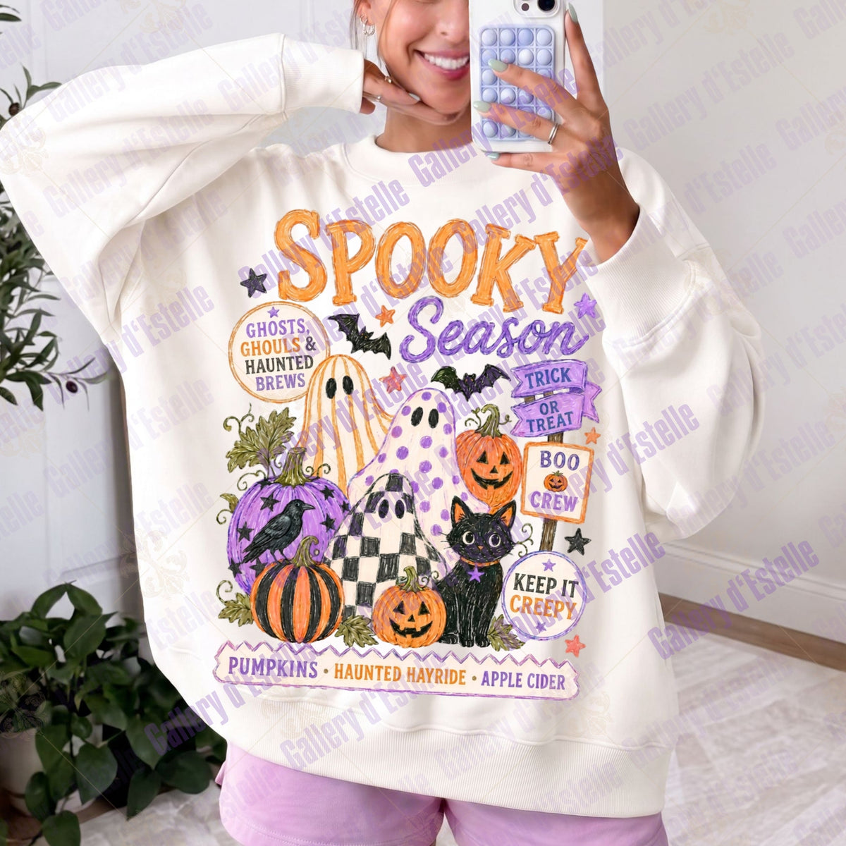 A person taking a mirror selfie wearing a cream-colored sweatshirt with a colorful 'Spooky Season' digital design featuring pumpkins, ghosts, a cat, and orange text, paired with light purple leggings.