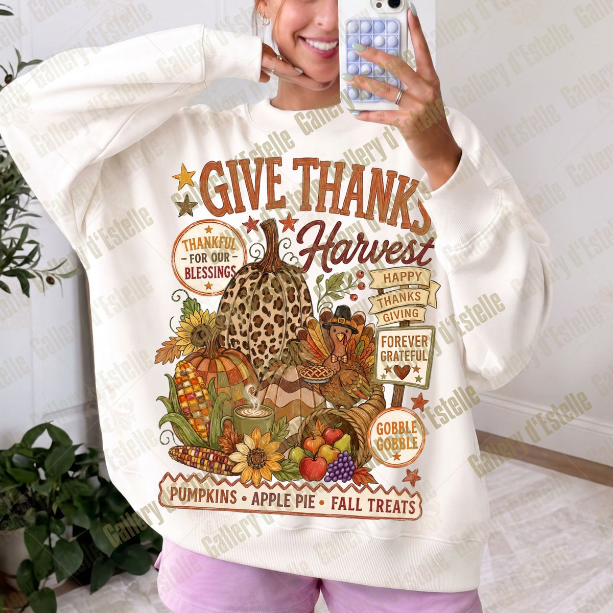 A woman takes a mirror selfie wearing a cream-colored sweatshirt with a large, colorful autumn harvest graphic on the front. The graphic features pumpkins, an apple pie, fall treats, and Thanksgiving-themed text.