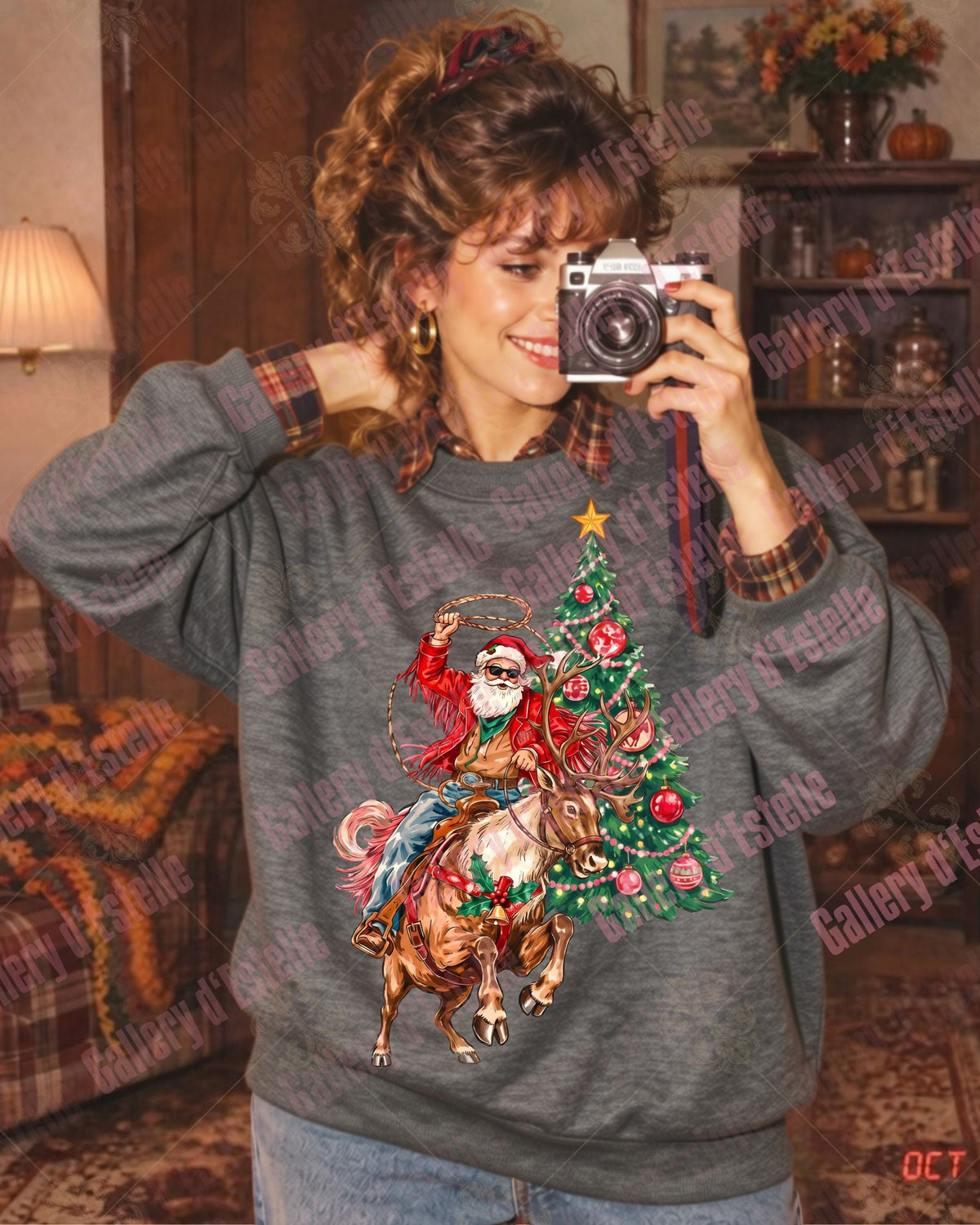 A woman takes a mirror selfie wearing a dark gray crewneck sweatshirt with a graphic of a Santa-clad cowboy riding a bucking reindeer beside a decorated Christmas tree. The photo is taken in a cozy, vintage-style living room with a lamp, plaid armchair, and wooden bookshelf.