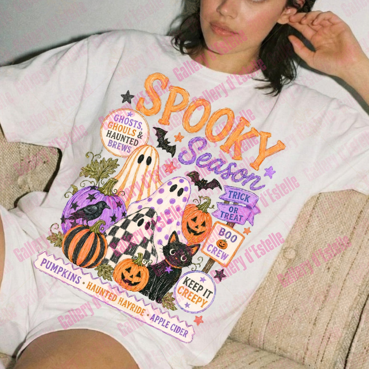 A woman reclining on a beige couch wearing a white graphic T-shirt with a colorful Halloween-themed print that reads 'Spooky Season' and includes pumpkins, ghosts, bats, a cat, and autumn phrases.