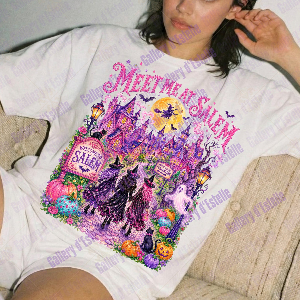 A woman reclining on a beige couch wearing a white graphic t-shirt with a colorful Halloween-themed print that reads 'MEET ME AT SALEM'