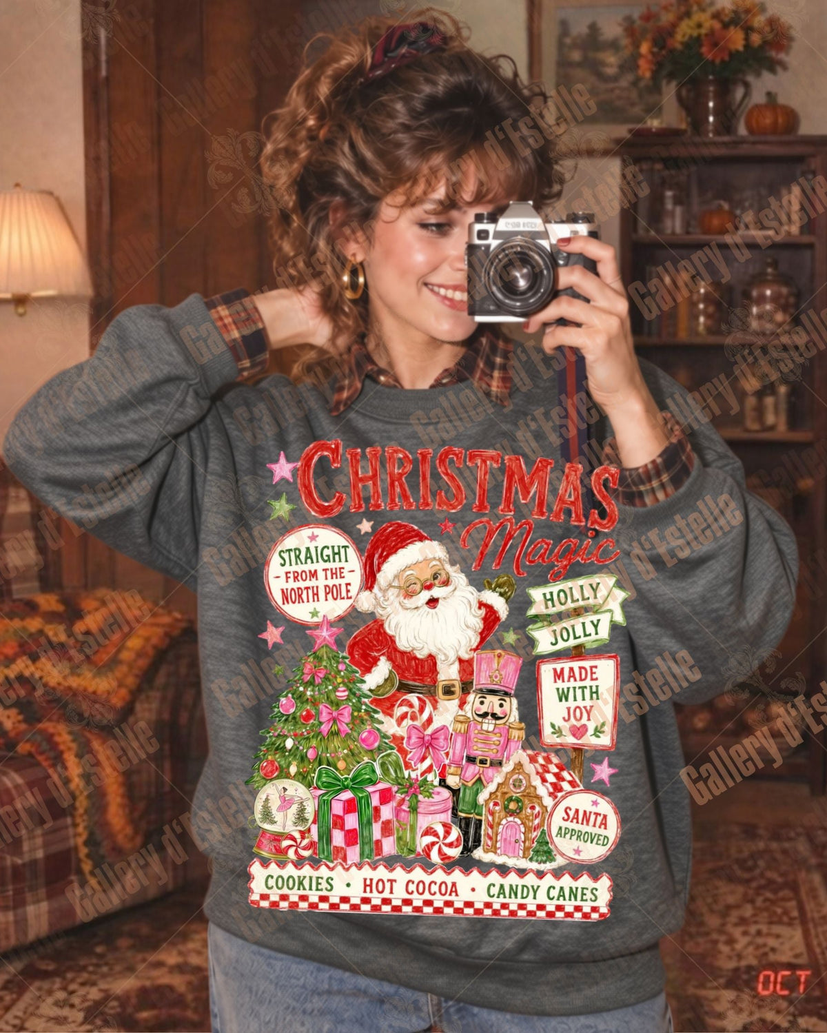 A woman takes a mirror selfie wearing a gray long-sleeve crewneck sweatshirt with a colorful vintage Christmas graphic featuring Santa Claus, candy canes, ornaments, and festive text. The photo is taken in a cozy, retro-styled living room with a plaid armchair and warm lighting.