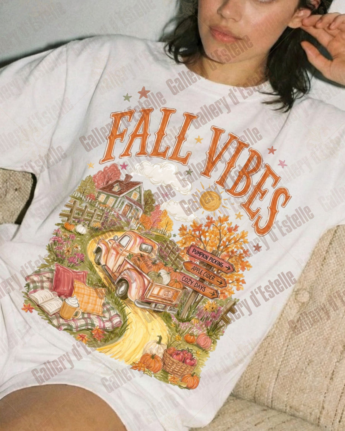 A woman reclining on a beige couch wearing a white graphic T-shirt with orange 'FALL VIBES' text and a colorful autumn scene print, paired with white shorts.