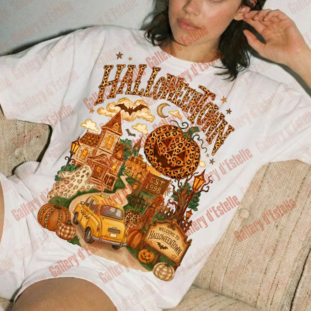 A woman reclining on a beige couch wearing a white graphic t-shirt with a Halloween-themed print featuring a haunted house, pumpkin, yellow truck, and the word 'HALLOWEEN' in orange. 