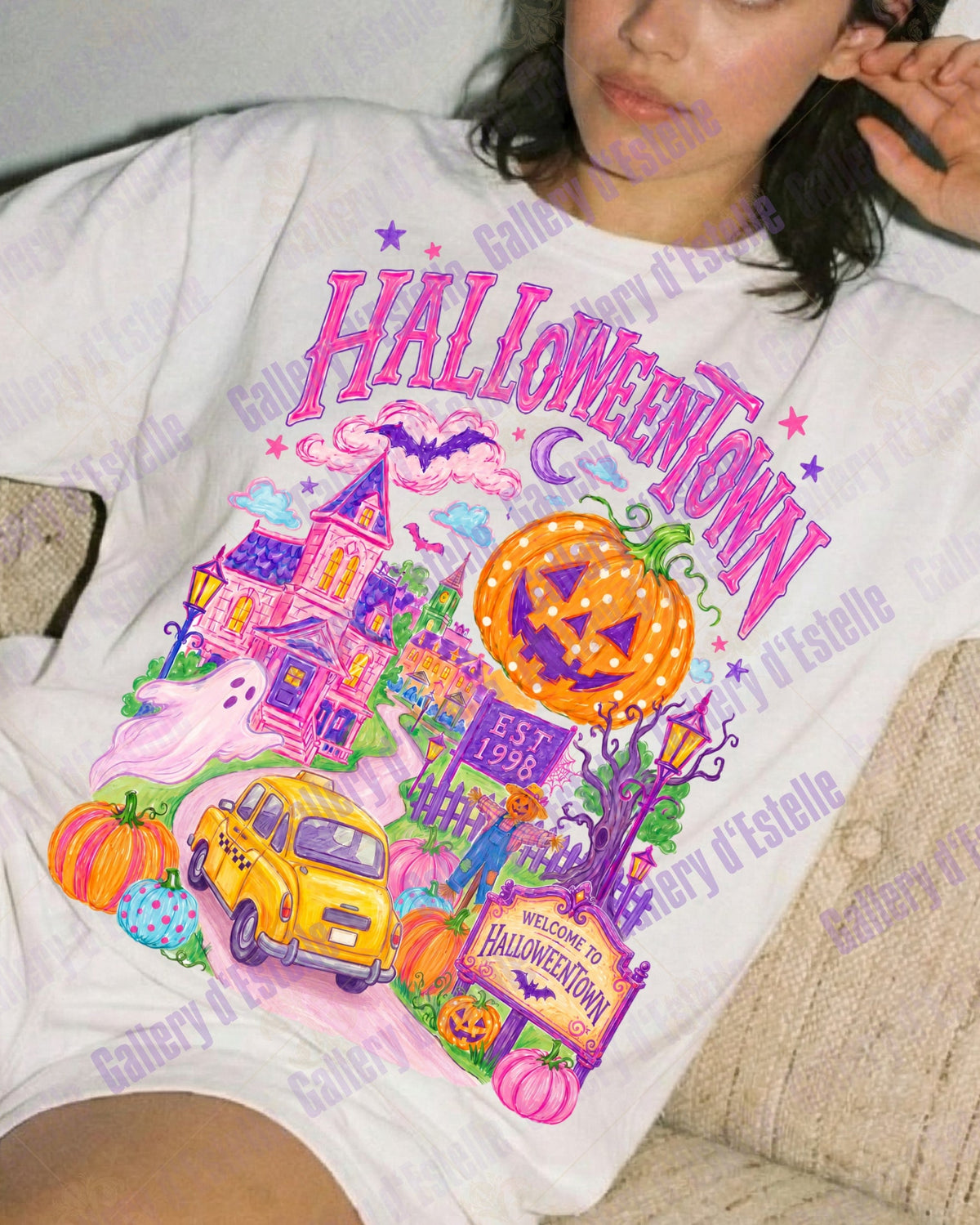 A woman reclining on a beige couch wearing a white graphic t-shirt with a colorful Halloween-themed print that reads 'HALLOWEENTOWN' above a haunted house, pumpkin, car, and ghost illustration.