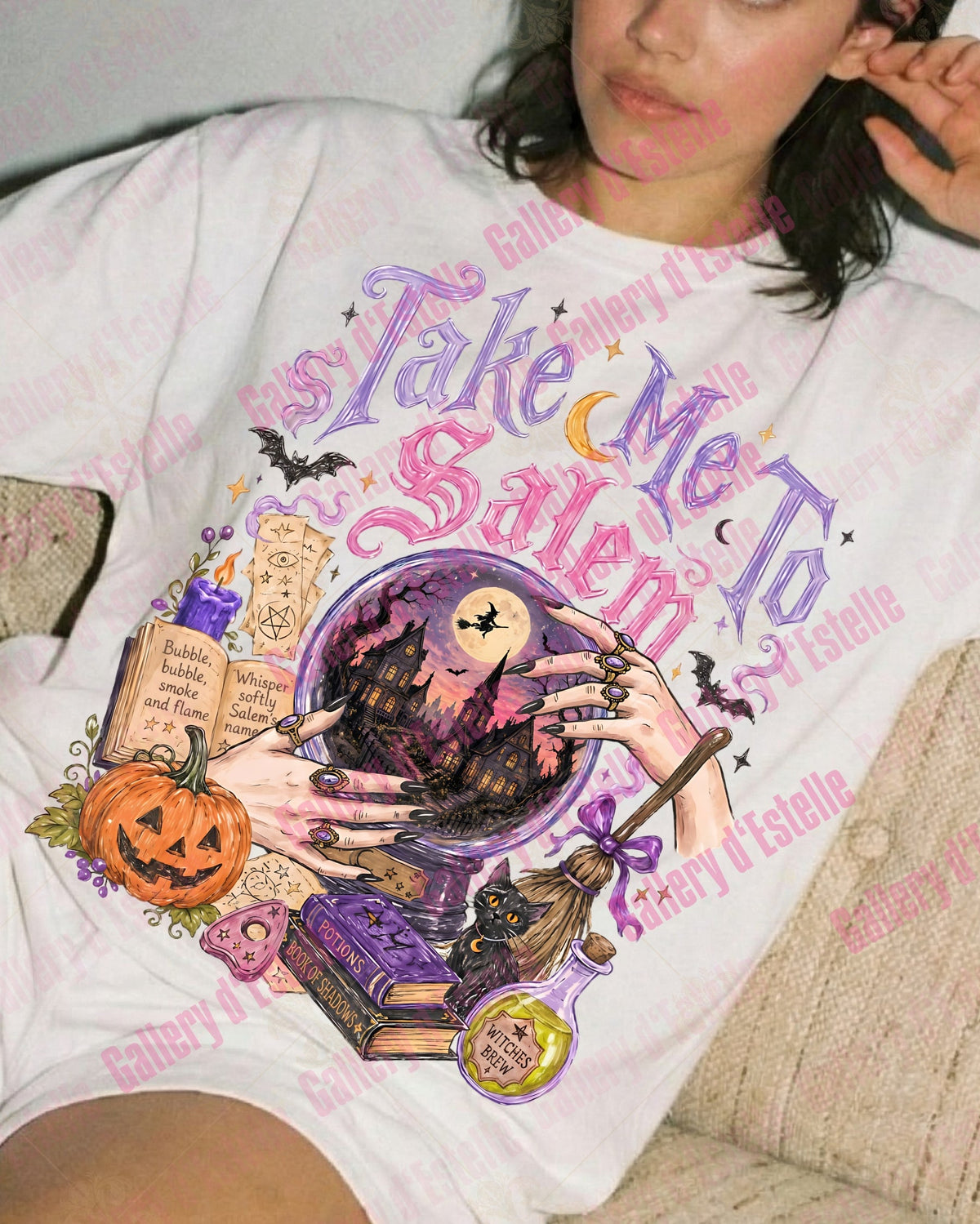 A woman reclining on a beige couch wearing a white graphic T-shirt with a Halloween-themed illustration and the text 'Take Me To Salem.' The shirt features a witch, pumpkin, book, broom, and bats in purple, orange, black, and pink tones.