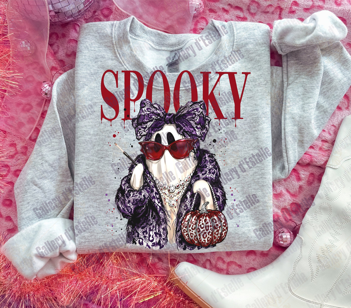 A light gray sweatshirt laid flat on a pink textured surface, featuring a graphic of a ghost in a purple leopard-print outfit with red sunglasses and the word 'SPOOKY' in red above it. White boots and sheer pink fabric are styled around it.