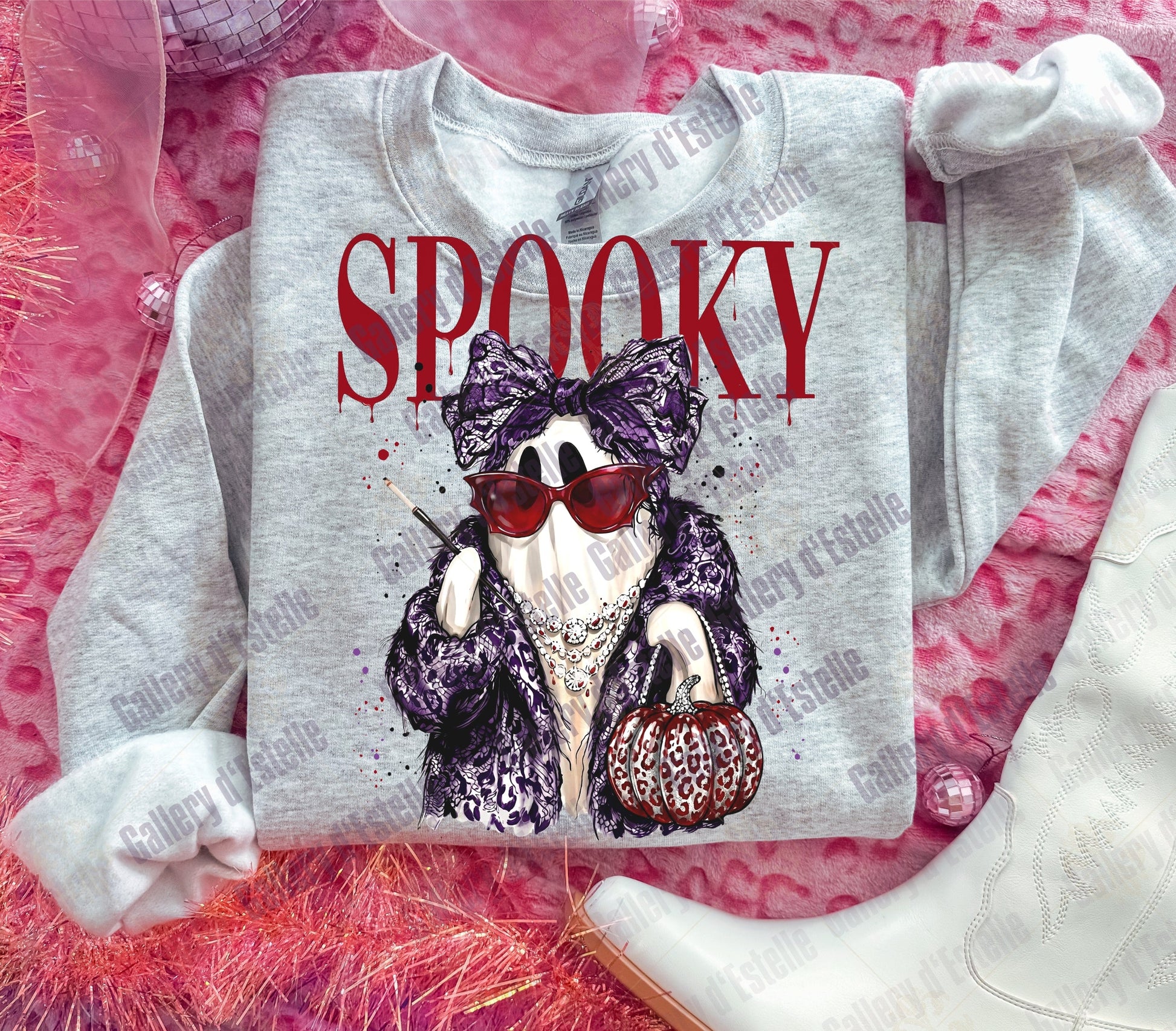 A light gray sweatshirt laid flat on a pink textured surface, featuring a graphic of a ghost in a purple leopard-print outfit with red sunglasses and the word 'SPOOKY' in red above it. White boots and sheer pink fabric are styled around it.