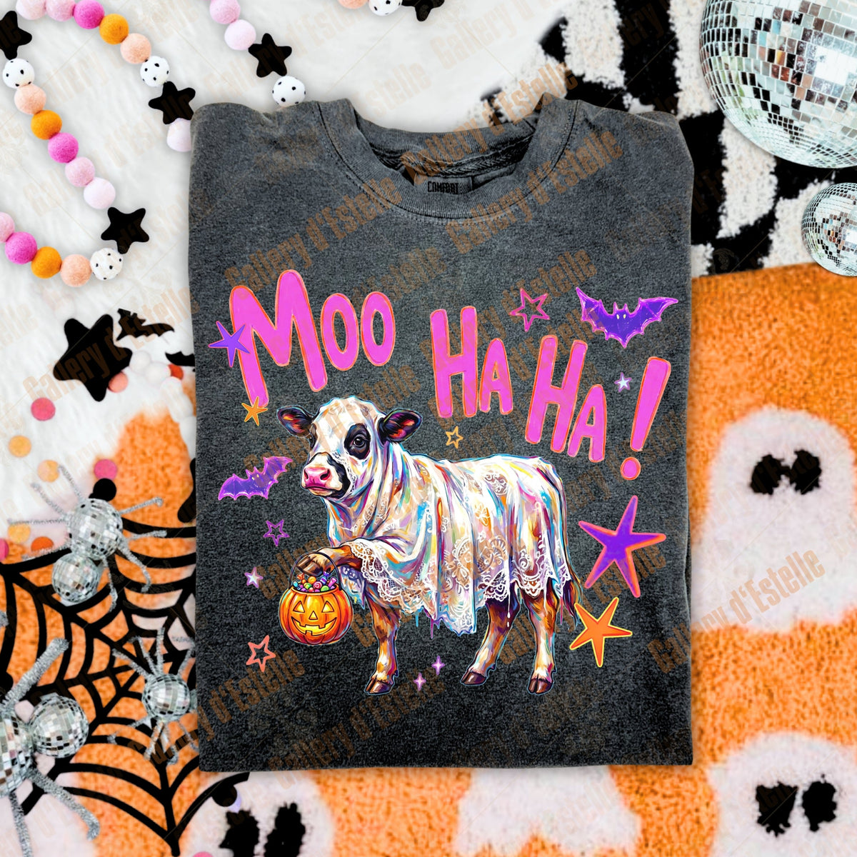 A folded dark gray t-shirt with a colorful Halloween graphic featuring a cow in a lace dress, a jack-o'-lantern, and the text 'Moo Ha Ha!' in pink, displayed on a themed flat-lay background with spiderwebs, a disco ball, and ghost blankets.