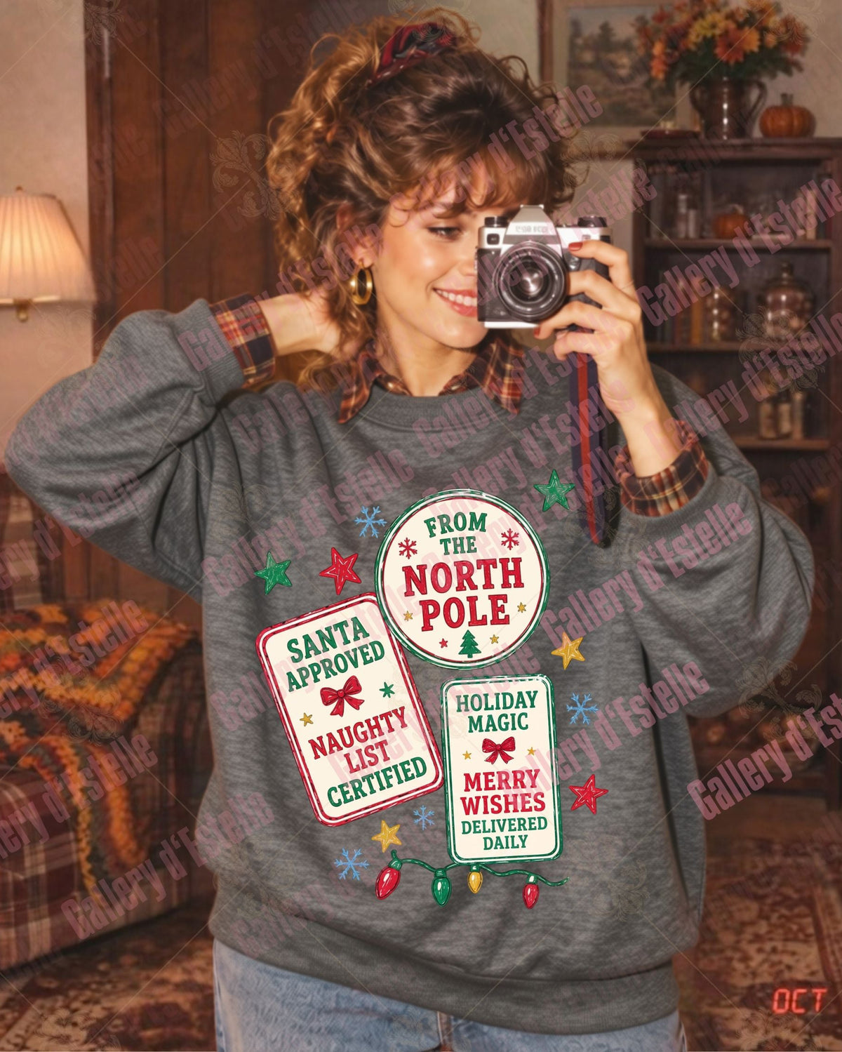 A woman takes a mirror selfie in a cozy living room, wearing a gray crewneck sweatshirt with a festive holiday graphic that reads 'From the North Pole' and 'Santa Approved'.