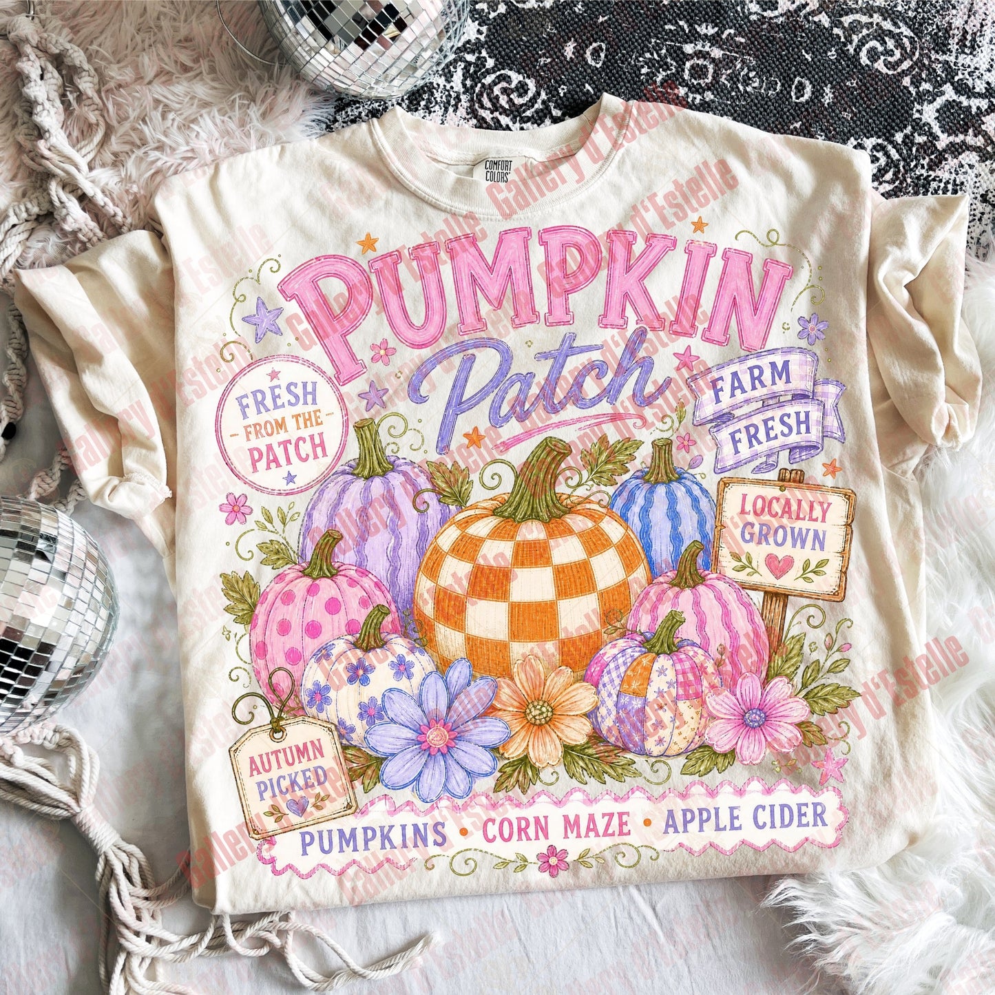 A cream-colored graphic t-shirt featuring a colorful autumn 'Pumpkin Patch' design with pumpkins, flowers, and seasonal text, styled in a flat-lay arrangement with a lace top, white faux fur, and a disco ball.