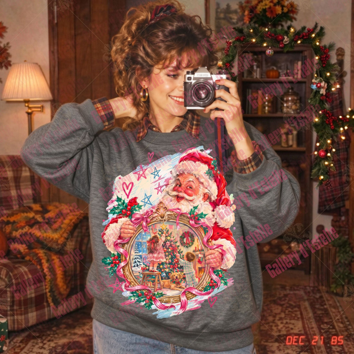 A woman takes a mirror selfie wearing a gray crewneck sweatshirt with a large, colorful Christmas graphic of Santa Claus holding an ornate golden picture frame depicting a nostalgic Christmas scene, with two children decorating a beautifully lit Christmas tree alongside their adorable dog. The background shows a cozy, decorated living room with a Christmas tree and shelves.