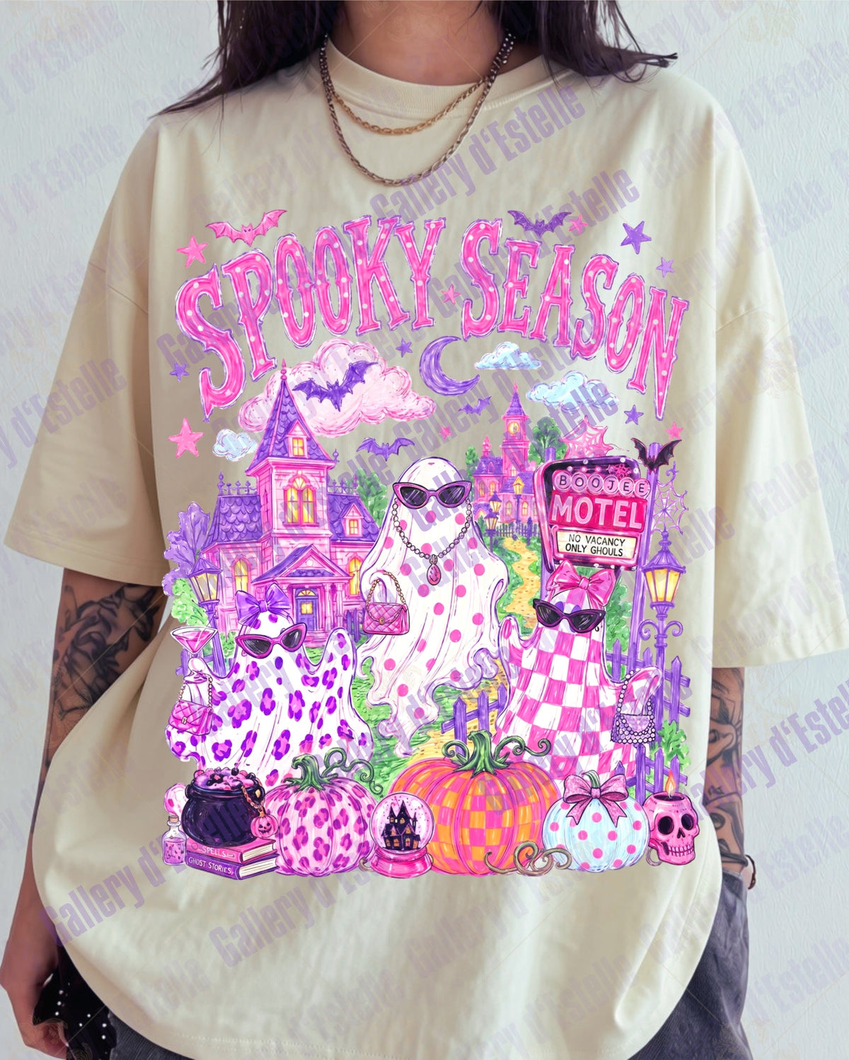 A person wearing an oversized cream-colored graphic t-shirt with a vibrant 'Spooky Season' Halloween print in pink, purple, and orange. The graphic features a haunted house, three ghosts, pumpkins, and a motel sign.