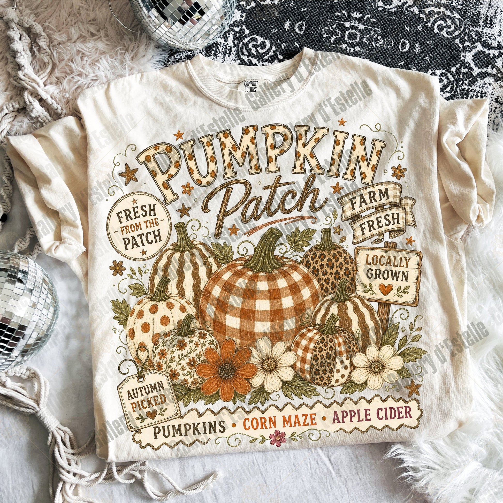 A cream-colored graphic t-shirt featuring a fall harvest design with pumpkins, flowers, and text reading 'Pumpkin Patch Farm Fresh' and 'Locally Grown'. The shirt is styled in a flat-lay arrangement with a black lace top, white faux fur, and a disco ball.