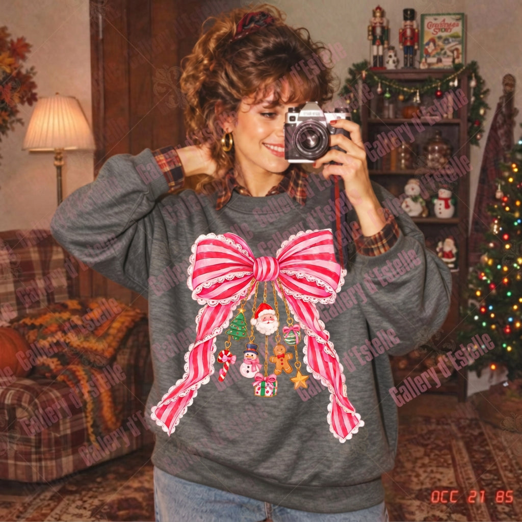 A woman taking a mirror selfie in a cozy, vintage-style living room, wearing a dark gray crewneck sweatshirt with a large pink and white striped bow graphic and a Christmas wreath design on the front.