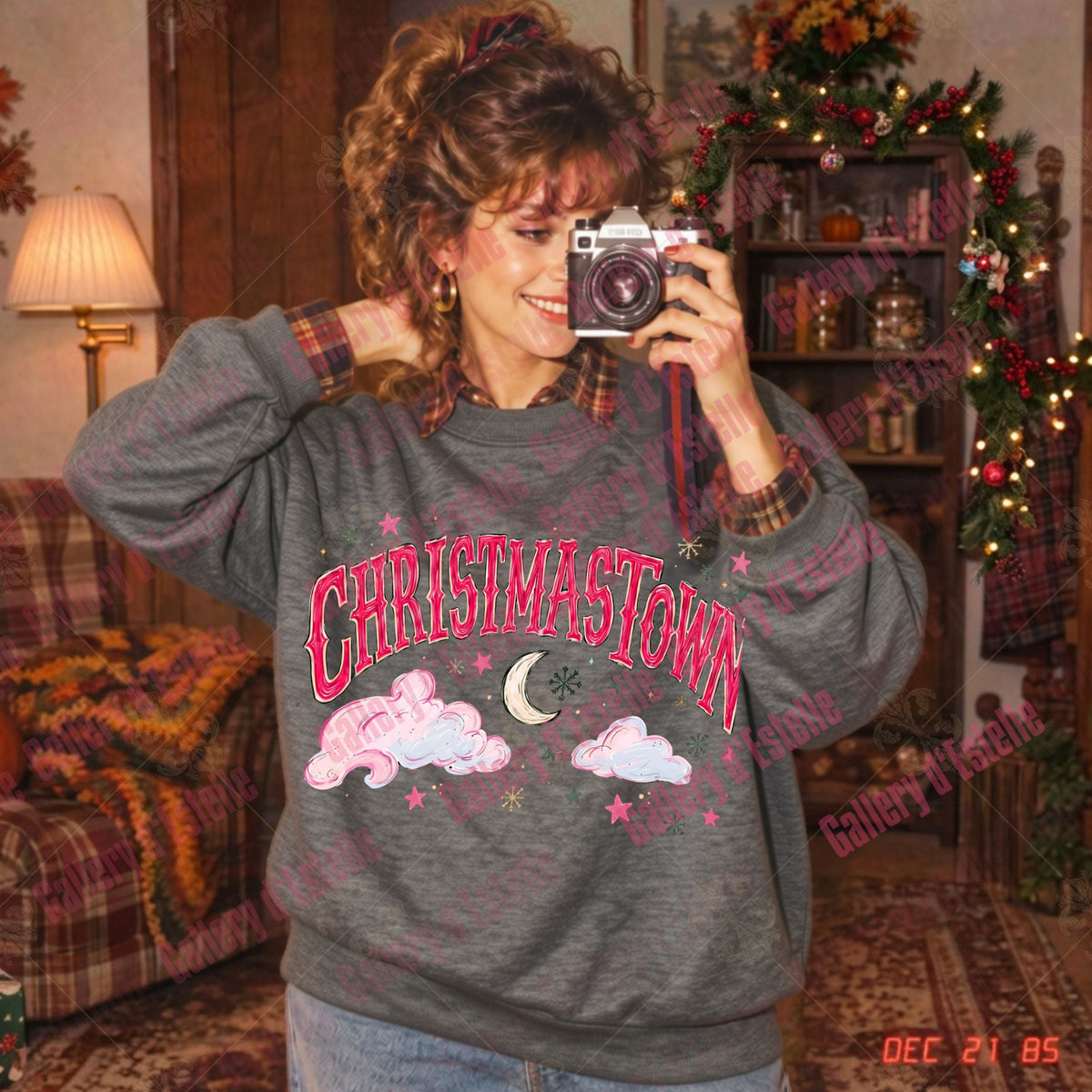A woman takes a mirror selfie wearing a dark gray crewneck sweatshirt with 'CHRISTAMASTOWN' in pink letters, decorated with pink clouds, a crescent moon, and stars. The background shows a cozy, decorated holiday living room with a Christmas tree and shelves.