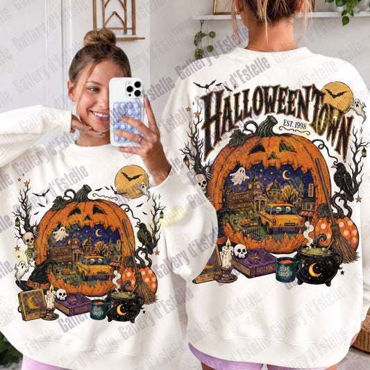 A person modeling a white sweatshirt shown from both front and back. The sweatshirt features a large Halloween-themed graphic with a jack-o'-lantern, haunted house scene, bats, cauldron, skulls, and the text 'HALLOWEENTOWN EST. 1998'