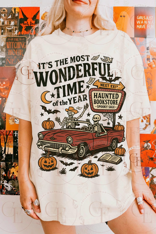 Person wearing a Halloween-themed t-shirt with graphics and the text ' It's the most Wonderful time of the year' featuring 2 skeletons driving a cara and a retro sign reading ' Haunted Bookstore '